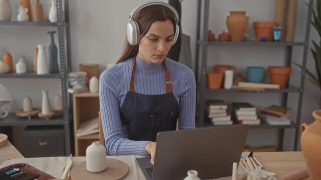 Woman in apron typing on laptop at artisan studio desk wearing headphones amid pottery and tools; concentration creativity.