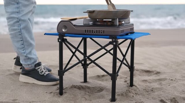 Camper cooking on portable stove on beach. Camper preparing meal on portable gas stove while at small folding table near ocean waves, enjoying outdoor cooking during beach vacation