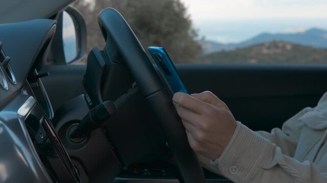 Woman using smartphone while driving modern car. Female driver gripping steering wheel, dangerously using smartphone while navigating winding mountain road during evening hours, risking accident
