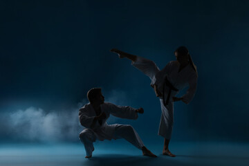 Obraz premium Man and woman in uniforms practicing karate on dark light blue background with smoke