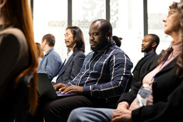 Obraz premium Black businessman takes notes on laptop at professional summit, forum conference with panel discussion and public speaker. Multiracial audience listen to ideas for entrepreneurship.
