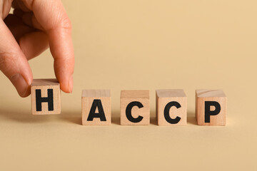 Cubes with abbreviation of HAZARD ANALYSIS AND CRITICAL POINTS and human hand on beige background