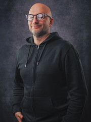 Fototapeta premium Man in his 40s or 50s wearing dark hoodie, grey painted background. Portrait of a male character with friendly face. Bald model with short grey beard and slim athletic body type and reading glasses.