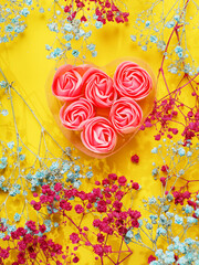 A heart made of pink flowers is surrounded by blue and pink Baby breath or Gypsophila flowers. The heart is in a clear container and is placed on a yellow background. Abstract floral design.