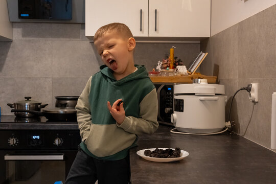 Funny little boy in green hoodie making disgusted face with tongue out while tasting dried prunes in modern kitchen at home. Concept of childhood, picky eater, healthy food and kids nutrition.