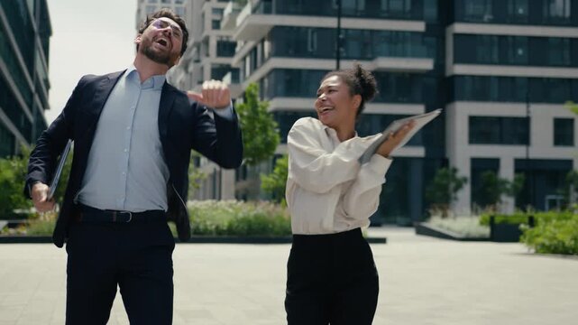 Two joyful active multiracial business colleagues dancing outside city celebrating success Caucasian businessman African businesswoman coworkers male female dance victory party corporate fun movement