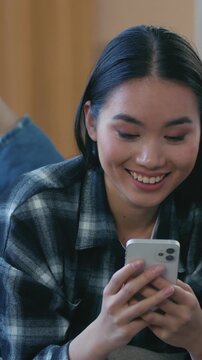 Asian woman lying on sofa at home smiling smartphone cellphone Korean Japanese female girl resting watching online video chatting on dating app texting ordering delivery relaxing indoors cozy leisure