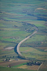 Obraz premium Long highway viaduct bending through green agricultural fields in western Sicily. The sweeping curve highlights modern infrastructure cutting across rural orchards and farmland on Mediterranean island
