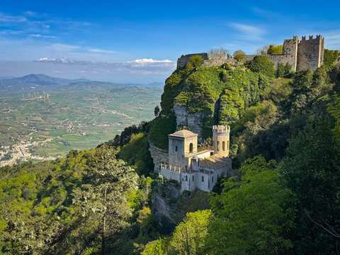 Stone fortress ruins perched on a lush cliff above a towered building and a wide Sicilian valley stretching into the distance. Picturesque view from Monte Erice, known for its charming historic town