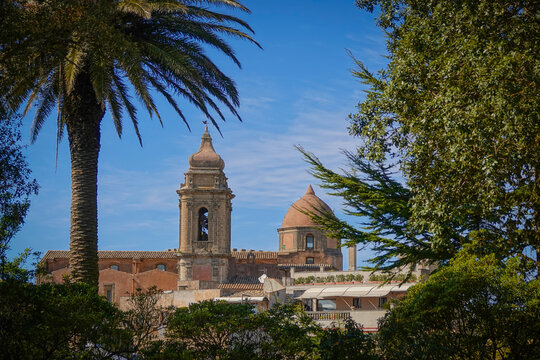 Lush palm tree and dense greenery frame the bell tower and domed church in old town of Erice in Sicily. Soft clouds streak a vivid blue sky that spreads above historic Sicilian sacral architecture.