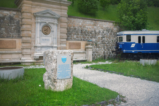 Semmering, Austria - May 28 2022: Carl Ritter von Ghega monument at Semmering railway station with memorial plaque and historic blue &Ouml;BB Class 5144 train in the background.
