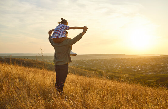 Happy father and child at sunset walking outdoors. Joyful shoulders ride in nature, arms raised across meadow as warm light paints hills, expressing trust, bonding, and joy. Fathers Day family love.