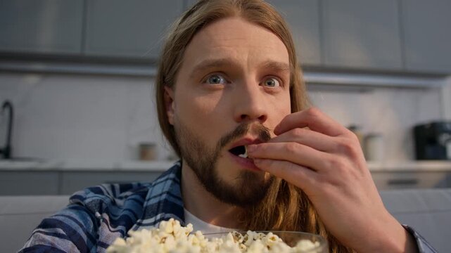 Caucasian man male guy eating popcorn while watching TV series at home weekend relaxing enjoying television film program media movie snack indoors leisure close up eat tasty corn entertainment hobby
