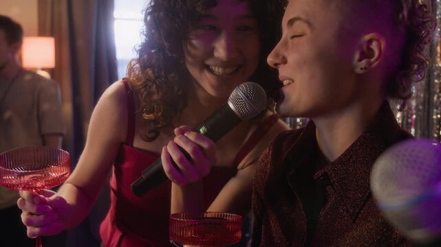 Tilt up view of two Caucasian fancy dressed girl friends holding red glass goblets singing karaoke at festive party with friends