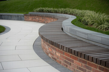Curved wooden bench without backrest mounted on a brick and concrete base lines a landscaped path in a sunny modern park.
