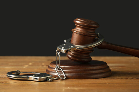 Handcuffs with judge's gavel on wooden table against dark background, closeup