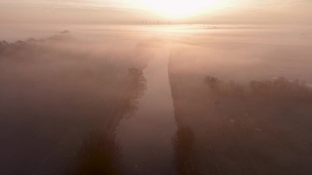 Aerial view of foggy canal at sunrise, flanked by trees and fields. Wind turbines silhouette the horizon as warm light diffuses through mist, evoking serenity, sustainability, and rural elegance.