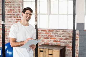 Man standing, writing on clipboard with pen in gym wearing white T-shirt by rack