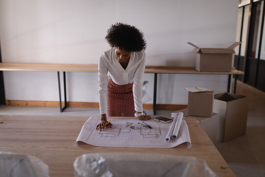 African American woman in blouse, print skirt leaning over studio table, marking plans with pencil