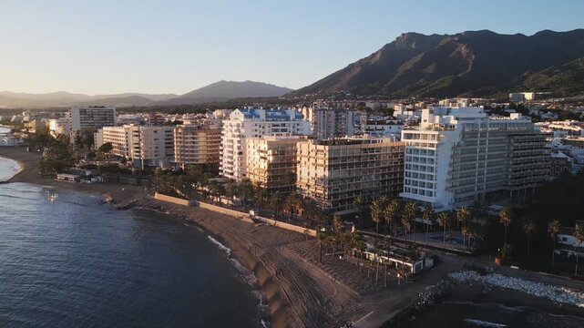 Beautiful wide drone shot tracking back along Marbella&rsquo;s coastline, glowing city skyline and luxury buildings with Sierra Blanca mountains in the background.