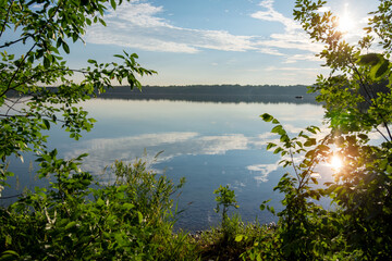 Framed by vibrant June foliage, the morning sun glistens over a calm Waukesha County lake. Serene Wisconsin summer landscape with blue sky.