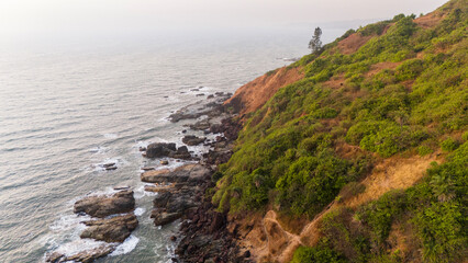 Obraz premium Aerial view of Arambol Beach, Goa, showing Arambol Mountain on the right with rugged rocks below as winter evening waves roll in from the left along the Arabian Sea coast.
