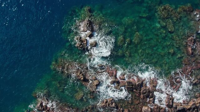 Top down drone orbit shot of turquoise sea waves hitting rocky coastline in slow motion. Cinematic water transparency and natural coral reef textures.
