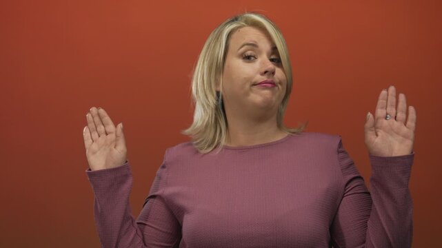 Young blonde woman holds up hands with palms forward in red studio with unimpressed expression; indifference.
