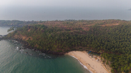Aerial drone view of Cabo de Rama Beach in Canaguinim, South Goa, showing white sand coastline, bluish ocean water, waves, rocky cliffs in the background, and abundant coconut palm trees