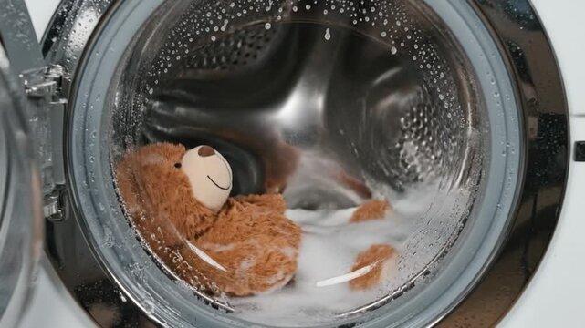 A brown teddy bear rotates in a washing machine with suds, viewed through the glass door