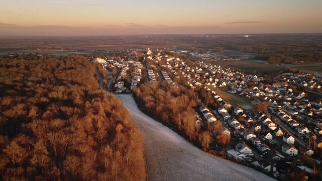 Forward-moving drone shot gliding along a forest edge over a frost-covered field, revealing Elchingen&rsquo;s village rooftops and surrounding countryside in warm golden-hour light.