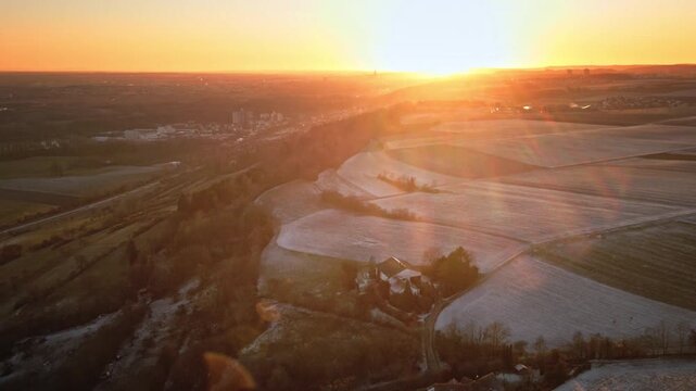 Drone pulls back to reveal a wide panorama of frost-covered farmland at sunrise, with warm golden glow, soft haze, and a farmhouse and field roads appearing below.