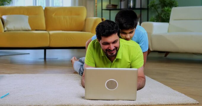 Indian asian man with son using laptop lying on carpet in modern living room, young father relaxing with child on back while browsing internet on computer at home lifestyle