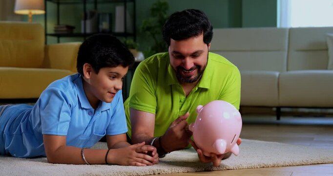 Indian asian young father son taking out coins from piggy bank with happiness while lying on carpet together, learning saving habits and enjoying joyful family bonding time in modern living room