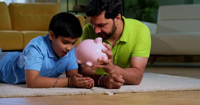 Indian asian young father son taking out coins from piggy bank with happiness while lying on carpet together, learning saving habits and enjoying joyful family bonding time in modern living room