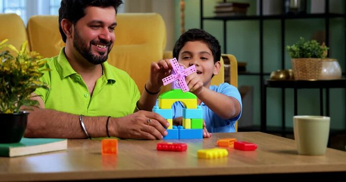 Indian asian young father son building toy windmill together in playful atmosphere, creating colorful plastic tower while spending quality family time in modern living room at home