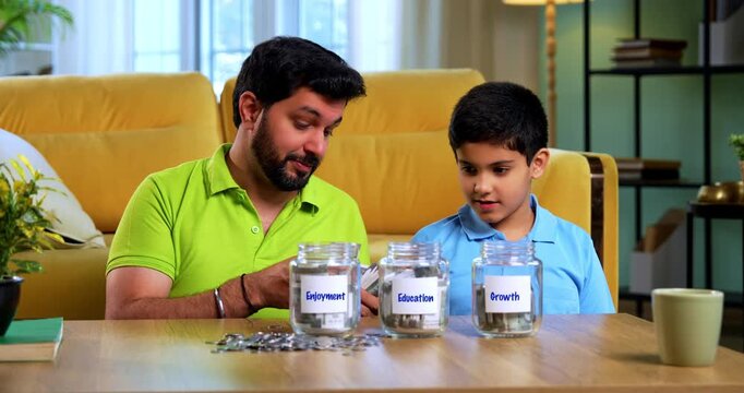 Indian asian young father teaching son importance of saving for growth, education and enjoyment using different labeled money jars while sitting together in modern living room at home