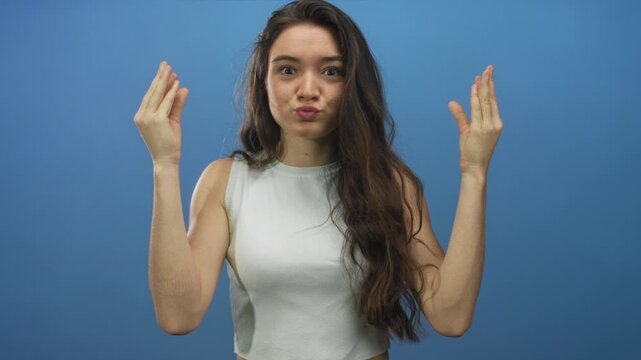 Teenage woman with puffed cheeks holds her temples while raising both hands in a studio with blue backdrop; shock.