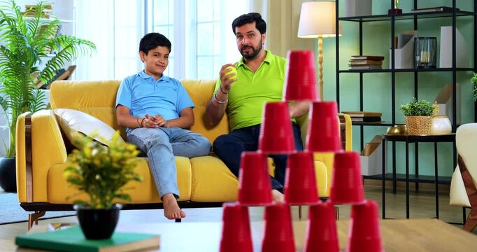 Indian asian young father son playing knock down stacked cups with ball game while lying on carpet and aiming plastic ball for fun activity together in modern living room at home