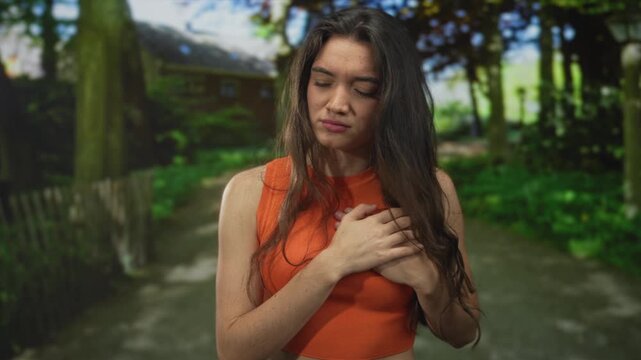 Young hispanic woman clutching bare chest and grimacing in a green park under dappled sunlight; discomfort.