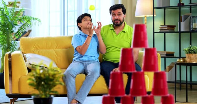 Indian asian young father son playing knock down stacked cups with ball game while lying on carpet and aiming plastic ball for fun activity together in modern living room at home