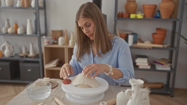 Blonde woman sculpts a ceramic vessel with her hands on a spinning pottery wheel in a studio; serenity.