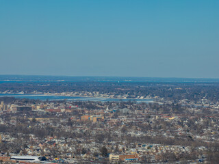 Urban landscape with snow dusted buildings and bare trees stretching towards a wide body of water and the distant horizon under a clear blue sky, capturing a cold weather cityscape