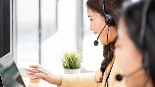 Happy Asian customer service agent wearing a headset and smiling while working with a colleague in a modern office.