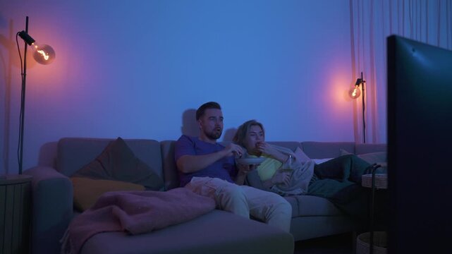 Couple watching TV at night while sharing a bowl of popcorn and wine on sofa
