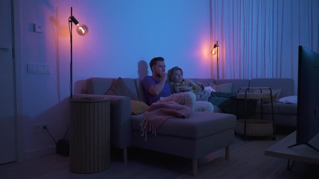 Couple watching TV at night while sharing a bowl of popcorn and wine on sofa