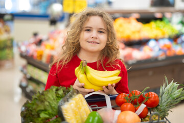 Child shopping with cart in supermarket. Kid choosing fresh vegetables in grocery store. Child with...