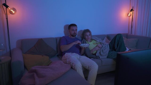 Couple watching TV at night while sharing a bowl of popcorn and wine on sofa