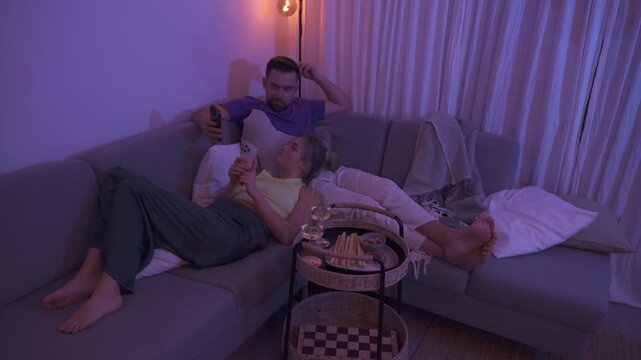 Couple Relaxing on Sofa at Night Using Smartphones with Snacks and Wine