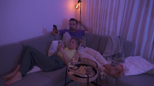 Couple Relaxing on Sofa at Night Using Smartphones with Snacks and Wine